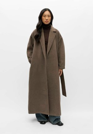 Femme portant un long manteau en laine marron sur un col roulé marron foncé et un jean à jambes larges, debout devant un fond blanc uni.