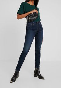 Žena v zeleném tričku a tmavých skinny jeans, drží černou ledvinku. Na nohou má stylové černé kotníkové boty a pózuje.