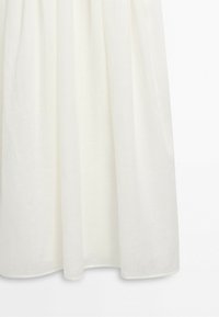 White gathered fabric hanging vertically with soft shadows on a plain light background, showing texture and flow details.