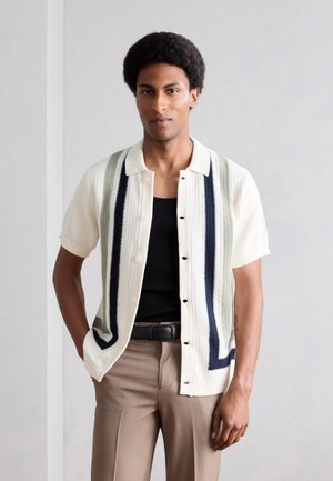 Junger Mann trägt ein weißes kurzärmeliges Strickhemd mit vertikalen schwarzen und grauen Streifen, ein schwarzes Tanktop und beige Hosen.