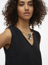 Göndör hajú nő fekete ujjatlan felsőt és ezüst virág alakú medál nyakláncot visel.