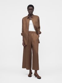 Black woman standing wearing brown wide-leg pants and matching button-up shirt over a white tank top with black heeled sandals.