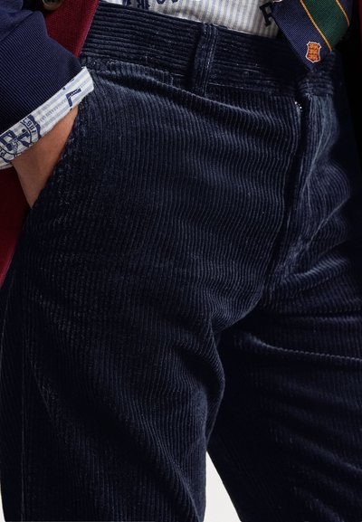 Marineblauwe corduroy broek met verticale ribbels, zijzakken en een knoopsluiting; textuur van de stof zichtbaar met een subtiele glans.