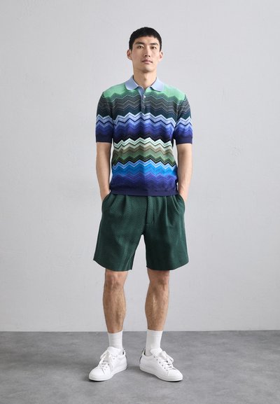 Polo cu model în zigzag multicolor, în nuanțe de albastru, verde și mov, asortat cu pantaloni scurți în herringbone verzi și adidași albi.