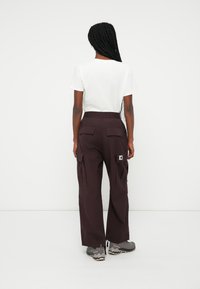 Pantaloni cargo bordeaux con vita elastica, dotati di due tasche laterali e un patch con logo. Abbinati a una maglietta bianca semplice.