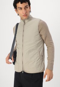 Beige gepolsterte Weste mit hohem Kragen, einem Reißverschluss vorne und seitlichen Taschen. Weiche, strukturierte Ärmel aus einem kontrastierenden hellbraunen Strick.
