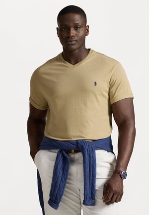 Khaki V-nyakú póló feltekert ujjal, fehér nadrággal és a derekon megkötött kék kötött pulóverrel párosítva. Az outfit része egy karóra is.