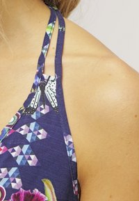 Top morado tipo halter con un patrón geométrico y floral, con tirantes estrechos y tela texturizada, que revela detalles en los hombros.