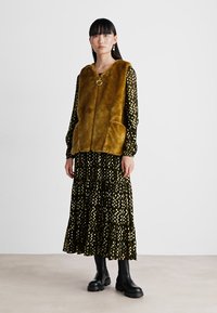 Faux-Fur-Weste in Olivgrün mit einem Frontverschluss und Taschen über einem schwarzen Maxikleid mit goldenen Polka-Punkten und gestuften Säumen, kombiniert mit schwarzen Knöchelstiefeln.