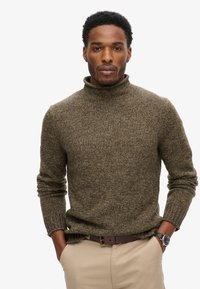 Bruine, gestructureerde coltrui met een slim fit ontwerp. Model draagt het met beige broeken en een horloge. De trui heeft lange mouwen en geribde manchetten.