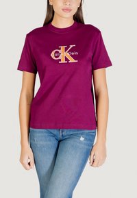 Camiseta de algodón color granate con un logo de Calvin Klein impreso en dorado y blanco. Manga corta, cuello redondo y un ajuste relajado. Combinada con pantalones vaqueros.
