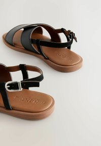 Sandales noires à lanières avec un mélange de textures lisses et en daim, une boucle ajustable et une semelle en caoutchouc marron comportant un logo en relief.