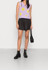 Lavendel mouwloze trui met een gele smiley-gezichtenpatroon, gecombineerd met een zwarte geplooide mini-jurk en zwarte enkellaarsjes. Zwarte tas inbegrepen.