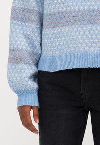 Pull en tricot bleu clair présentant un motif texturé avec des accents crème et marron, des poignets côtelés et une longueur cropped, associé à un jean noir.