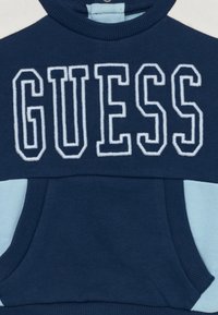 Sweat-shirt bleu marine avec des accents bleu clair et une poche frontale, arborant "GUESS" brodé en lettres blanches sur la poitrine.