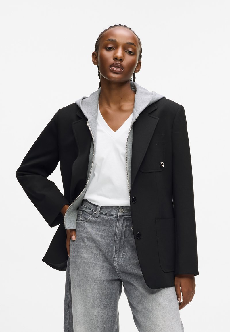 Čierny blejzer s šedou kapucňou, bielym V-ksichtovým tričkom a šedými džínami. Blejzer má vrecko a gombíkové uzávery, s hladkou textúrou.