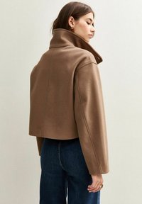 Kvinna i en kort brun kappa med hög krage, iförd mörkblå jeans, som står sidan mot en enkel ljus vägg.