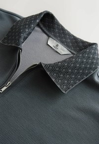 Mørkegrå ribstrikket skjorte med en krave med geometrisk mønster og en delvist åbnet sølvlynlås, mærket "NEXT" inde i kraven.