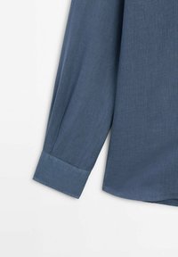 Blå långärmad skjorta med en mjuk textur, knäppning vid ärmslut och rak nederkant. Tyget verkar lätt och har ett subtilt väv-mönster.