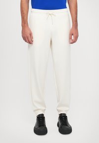 Pantalons de survêtement blancs en tissu doux, avec une taille à cordon de serrage et un design fuselé. Portés avec des baskets noires.