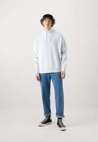 Lichtgrijze hoodie met een trekkoord, ontspannen pasvorm, kangoeroezak, gecombineerd met blauwe jeans en zwarte hoge sneakers.