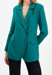 Blazer bleu sarcelle à coupe ajustée, col à revers crantés, fermeture à un bouton et deux poches avant. Tissu lisse avec une finition élégante.