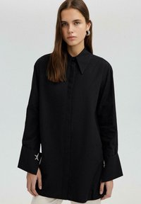 Black shirt with long sleeves and a pointed collar. Features silver star-shaped accents on cuffs. Made of a smooth fabric with a relaxed fit.