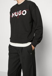 Sudadera negra con el texto "HUGO" en blanco y acentos rojos, cuello redondo, puños acanalados, superpuesta a una camisa blanca, combinada con pantalones negros.