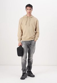 Sweatshirt bege com capuz, jeans cinzentos e sapatilhas de desporto pretas. Modelo segura um pequeno saco preto. Design simples, textura suave, ajuste casual.