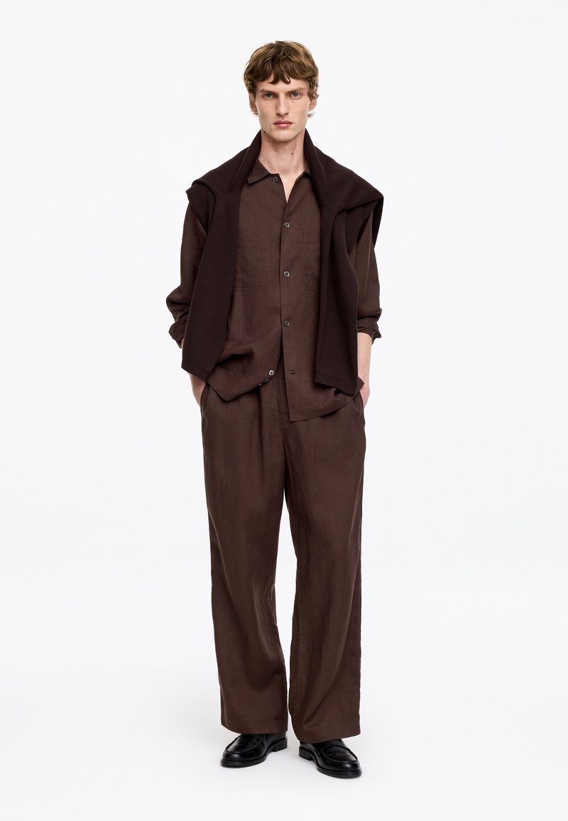 Jeune homme portant une chemise ample marron foncé et un pantalon, avec un pull sombre drapé sur les épaules et des chaussures noires, debout les mains dans les poches.
