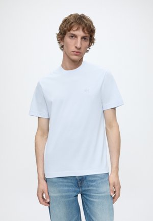 Giovane uomo con capelli ricci che indossa una semplice maglietta bianca a maniche corte e jeans blu, in piedi davanti a uno sfondo bianco.