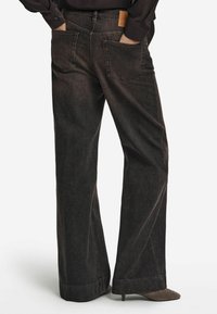 Jean noir à jambes larges en denim, avec une taille haute, un design à cinq poches et une étiquette de marque en cuir marron à l'arrière.