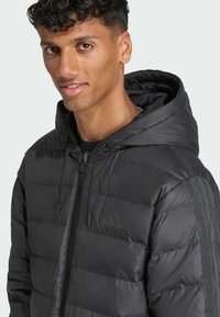 Czarna puchowa kurtka z kapturem, z pikowanymi sekcjami, sznurkami oraz subtelnym logo na piersi. Teksturowany materiał o gładkim wykończeniu.