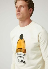 Sudadera de color crema con un gráfico de un gorro naranja con un pompón verde, con texto impreso debajo. Tela suave y cuello de canalé.