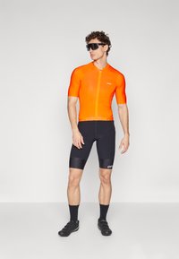 Pánska cyklistická dres s krátkym rukávom v jasne oranžovej farbe s predným zipsom, kombinovaný s čiernymi textúrovanými šortkami a čiernymi cyklistickými topánkami.