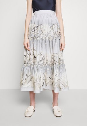 Femme portant une jupe midi à volants gris clair avec un imprimé coquillages et thème océan, associée à des chaussures blanches slip-on.