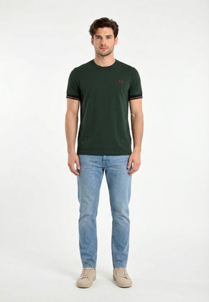 Giovane uomo in piedi, frontale, indossa una maglietta verde scuro, jeans azzurri e sneakers beige su uno sfondo bianco uniforme.