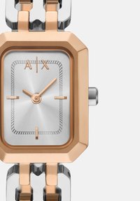 Obdĺžnikové hodinky s púzdrom z ružového zlata a nerezovej ocele, strieborným ciferníkom a tromi hodinovými značkami. Kovový náramok s nesúvislými článkami v ružovom zlate a striebre.