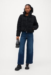 Svart cropped puffer jakke med kviltet tekstur og høy krage, kombinert med utsvingte blå jeans og kraftige svarte støvler, mens den holder en veske.