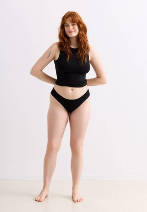 Rothaarige Frau steht barfuß, trägt ein schwarzes ärmelloses Oberteil und schwarze Unterwäsche, lächelt mit den Händen hinter dem Rücken.