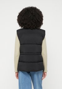 Eine Person mit lockigem Haar trägt eine schwarze Daunenweste über einem beigen gerippten Langarmshirt und lockeren blauen Jeans und ist mit dem Rücken zur Kamera gewandt.