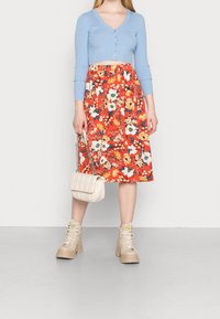 Ljusblå randig cardigan med V-ringning, röd midiklänning med blommönster, beige kängor och en krämfärgad kviltad handväska.