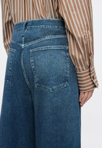 Sinised teksapüksid kõrge vöökohaga ja laiade säärtega, millel on tagataskud ja nähtavad õmblused. Kandatud triibulise särgiga.