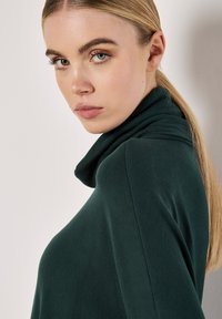 Tmavě zelený turtleneck z látky s měkkým, texturovaným povrchem. Obsahuje vysoký límec a elegantní design, na modelce s dlouhými vlasy.