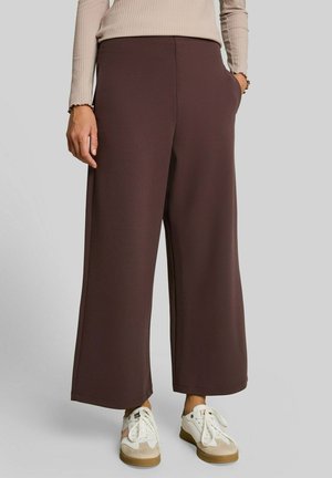 Frau trägt braune, weit geschnittene Hose, beige geripptes Langarmshirt und weiße Sneakers mit beigen und braunen Details.