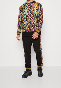 Färggad mönstrad sweatshirt med svarta träningsbyxor, med abstrakta mönster i gult, lila och teal. Svarta sneakers med böjda ränder.