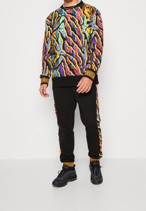 Sweatshirt coloré à motifs associé à un pantalon de survêtement noir, avec des designs abstraits en jaune, violet et teal. Baskets noires avec des rayures incurvées.