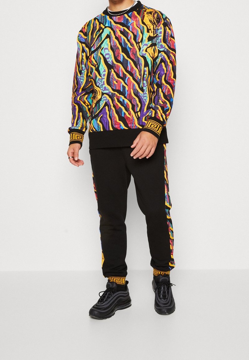 Färggad mönstrad sweatshirt med svarta träningsbyxor, med abstrakta mönster i gult, lila och teal. Svarta sneakers med böjda ränder.