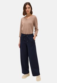 Béžový top s dlouhým rukávem v kombinaci s širokými námořnickými kalhotami. Kalhoty mají elastický pas a kapsy. Modelka má na sobě světlé tenisky.