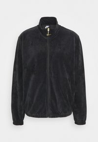 Černá kordurová bunda s vysokým límcem, zipem a elastickými manžetami. Na hrudi je decentní logo Nike a kontrastní zlaté kovové prvky.